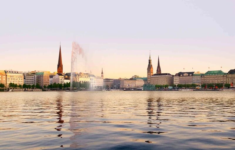 Blick &uuml;ber die Alster in Hamburg auf die Binnenalster-Promenade mit historischen Geb&auml;uden und Wasserfont&auml;ne bei Sonnenlicht